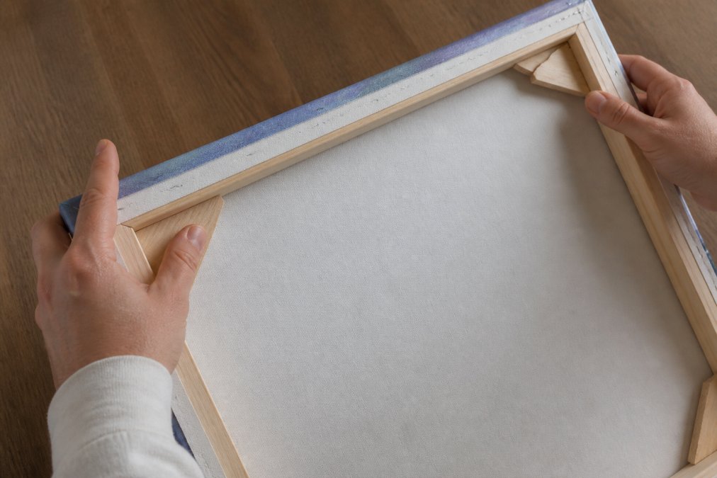 Hands examining the back of a stretched canvas, showing solid wood stretcher bars with visible joinery and corner reinforcement, wood grain texture clearly visible under studio lighting