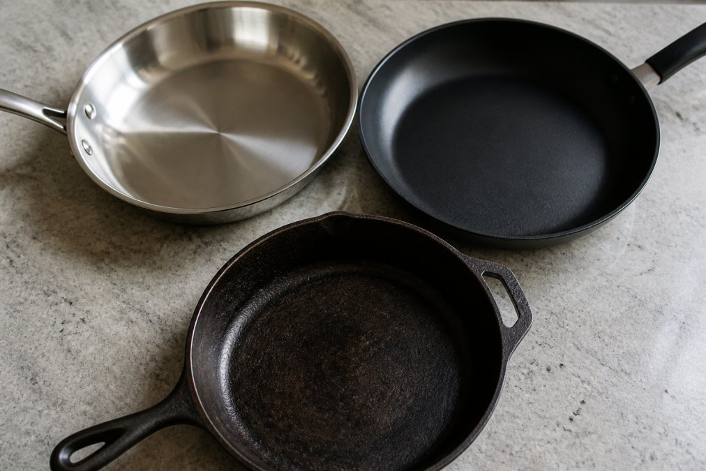 Close-up overhead shot of three different cookware pieces arranged on a kitchen counter with natural light highlighting their surfaces, a polished stainless steel pan with reflective finish, a dark nonstick skillet with matte coating, and a seasoned cast iron pan with deep patina, showing distinct material differences