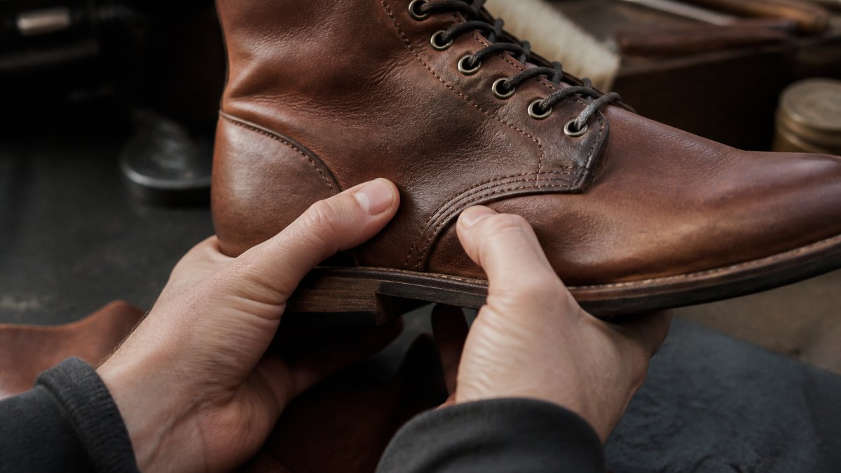 Close-up of hands examining premium full-grain leather boot, showing natural grain texture and subtle imperfections under natural daylight