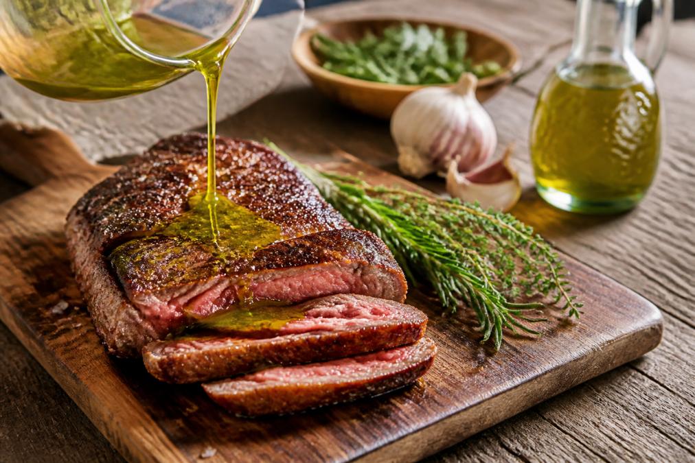 Golden-brown steak being drizzled with vibrant green olive oil on a wooden board with fresh herbs nearby