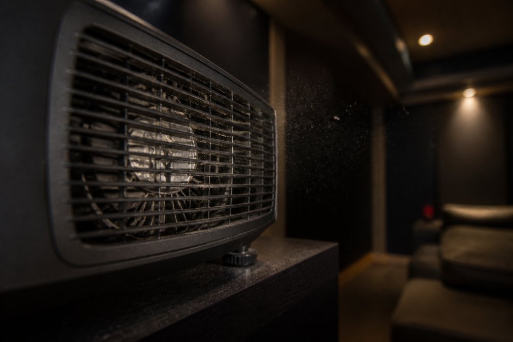 Close-up of a projector's ventilation grille and internal cooling fan with fine dust particles visible in the airflow, set in a dimly lit home theater room