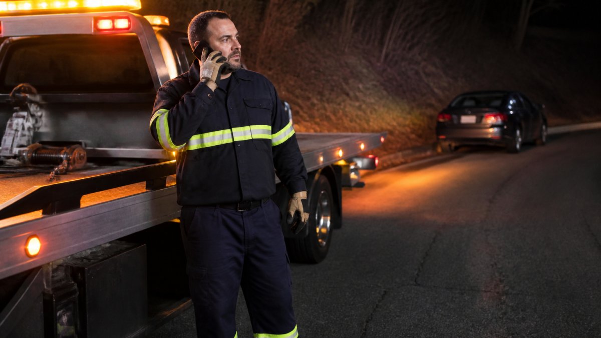 A tow truck operator speaking directly on a cell phone while standing beside a flatbed truck at night, with hazard lights glowing and a stranded car in the background