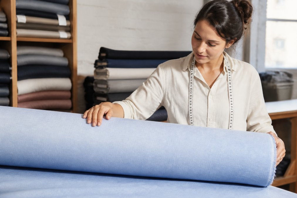 A professional seamstress examining a roll of poly-cotton blend fabric in a bright, organized textile studio, with rolls of fabric stacked neatly in the background