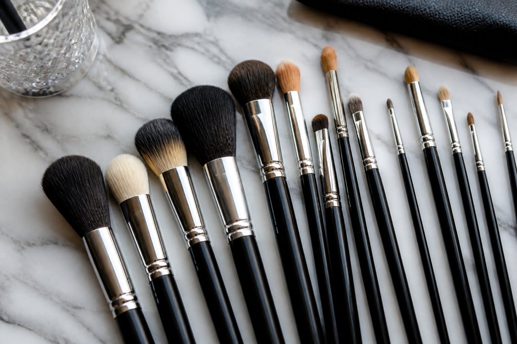 Close-up flat lay of a set of professional makeup brushes arranged neatly on a marble surface, showing varied shapes and bristle types in warm, natural lighting