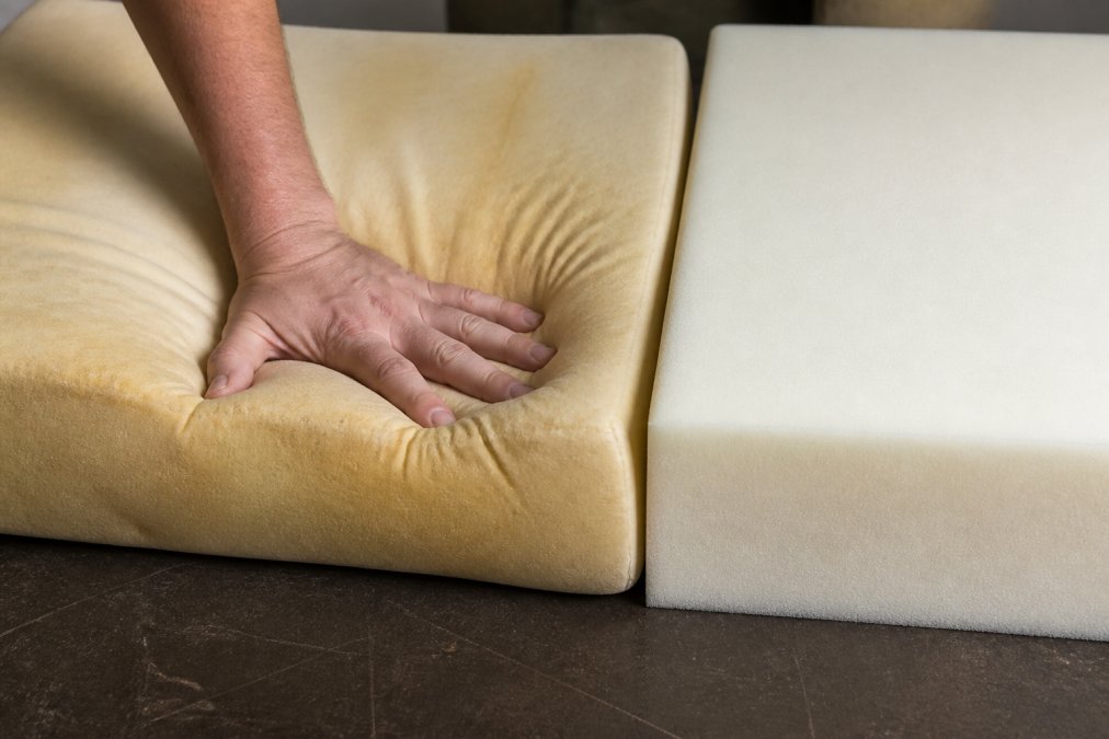 Close-up of a hand pressing into a worn foam cushion showing visible sagging and body impressions, with a newer dense foam sample beside it for comparison