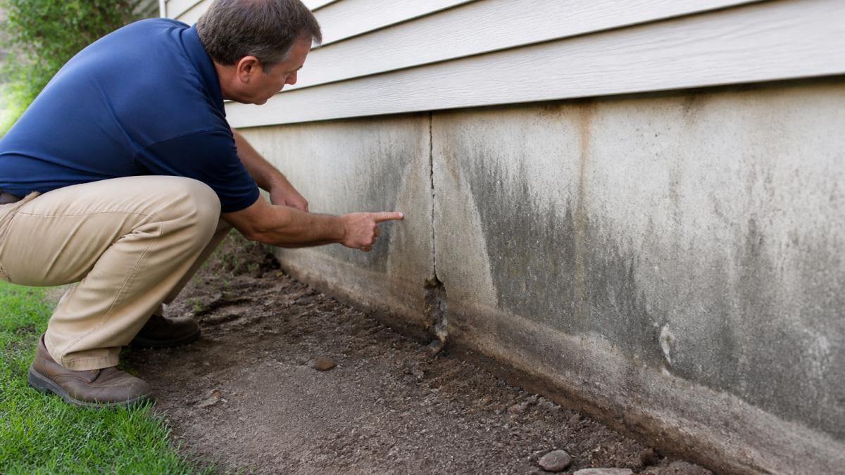 A homeowner crouching to inspect a visible crack in a concrete foundation wall with dark water staining and eroded soil along the base of a suburban house exterior