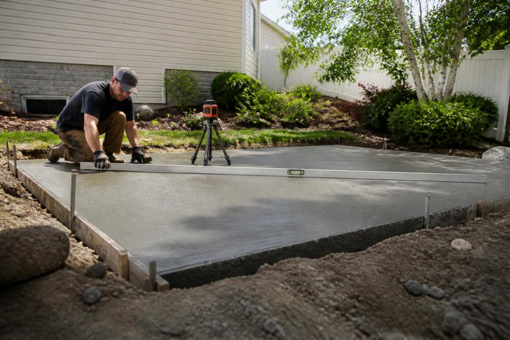 Professional installer measuring and leveling a concrete pad foundation for a hot tub installation, using laser levels and concrete finishing tools on a residential backyard site