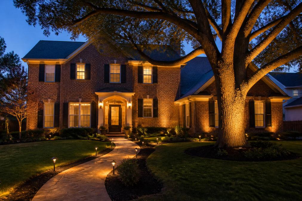 A two-story brick home at dusk with warm low-voltage lighting accenting the roofline, illuminating a stone walkway, and grazing light up the trunk of a mature oak tree