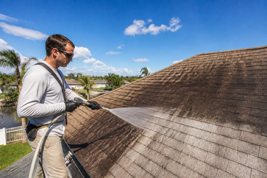 A technician in protective goggles and gloves applying a low-pressure soft wash solution to an asphalt shingle roof, black algae streaks visibly fading behind the spray nozzle, bright midday sun and blue sky