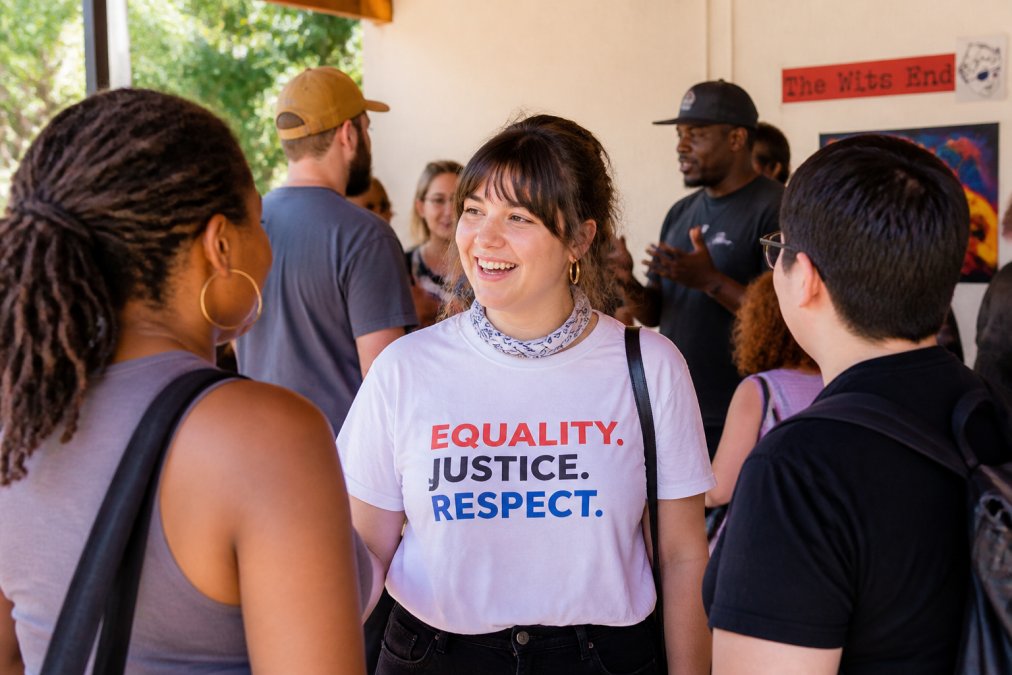 Person wearing a political statement t-shirt at a community gathering, standing among others engaged in conversation, natural outdoor lighting showing the apparel clearly