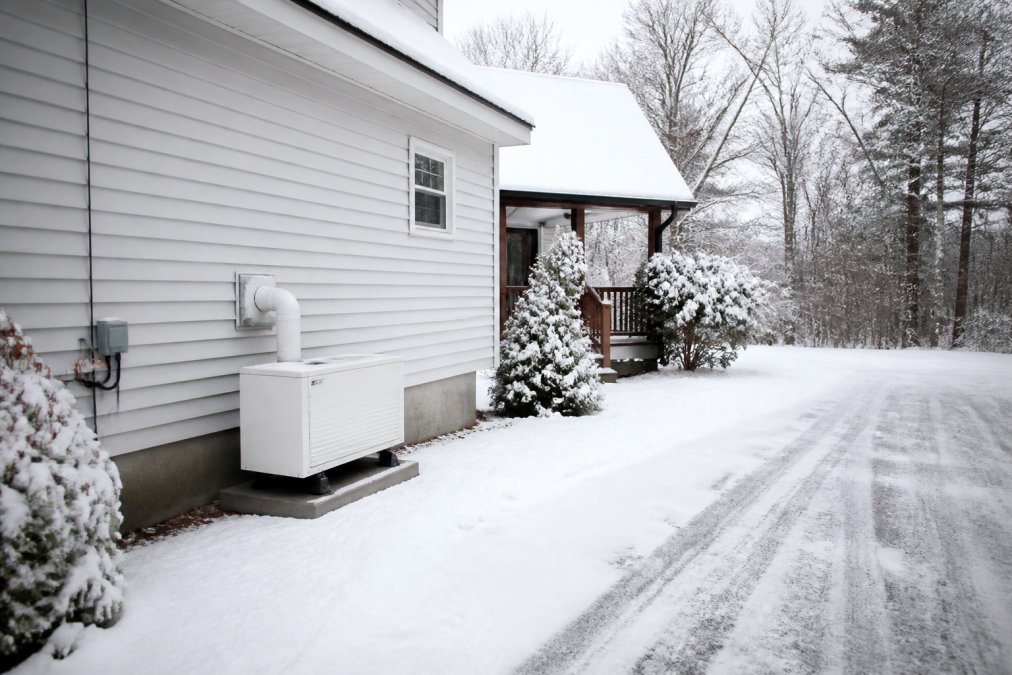Residential home in winter with snow covering the roof and driveway, heating system vent visible on the exterior wall, showing winter heating season demand reality
