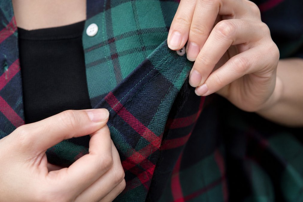 Close-up of hands examining fabric seams and stitching details on a quality plaid shirt, showing tight, even stitches and proper thread matching under natural daylight