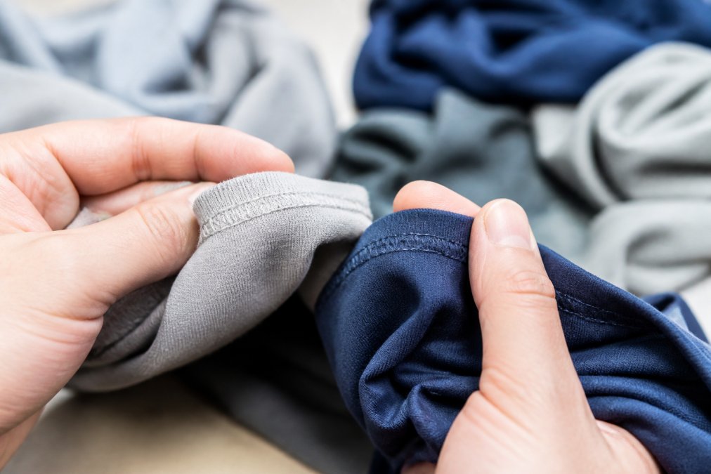 Hands carefully examining and comparing stitching quality on two different garments, showing the difference between dense, reinforced seams and loose, single-stitch construction under bright natural light