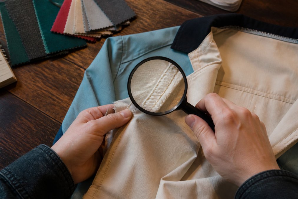 Close-up of a person's hands examining the interior seams and stitching of a vintage-style bowling shirt, with a magnifying glass and fabric swatches on a wooden table