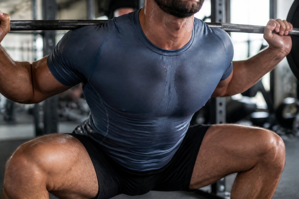 Close-up of athlete's torso during intense squat workout showing sweat-resistant technical fabric maintaining proper fit without riding up, demonstrating breathability and body-hugging support