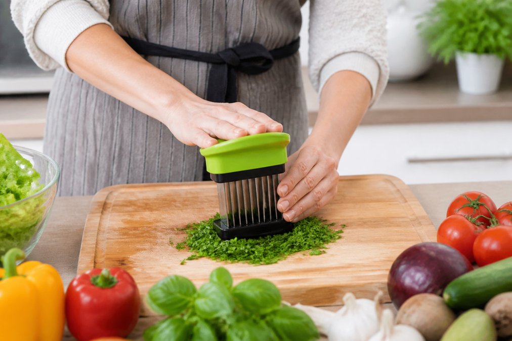 A home cook using a multi-blade kitchen tool to quickly chop fresh herbs on a wooden board in a bright, tidy kitchen with vegetables arranged nearby