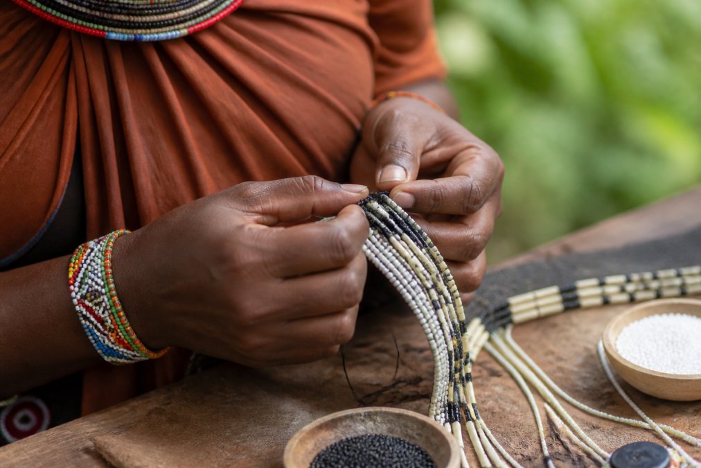 Artisan hands carefully weaving or beading a handmade accessory using traditional techniques, with natural fiber materials and tools visible, showing the intricate skill and cultural knowledge embedded in each piece