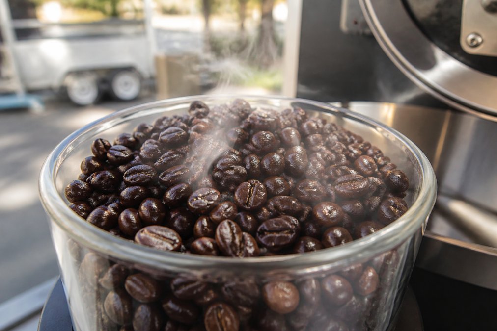 Close-up of freshly roasted coffee beans in a clear glass container showing glossy, dark surfaces with visible steam or vapor wisps rising from the beans in natural daylight