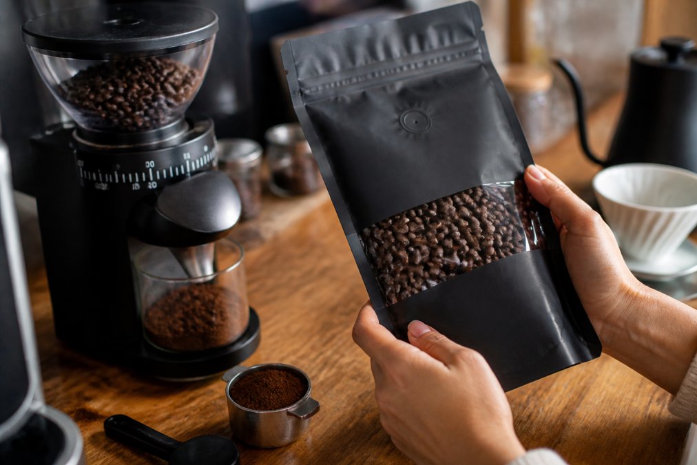 Hands holding a bag of roasted-to-order coffee with visible roast date label, with a burr grinder and freshly ground coffee visible in the background on a wooden countertop