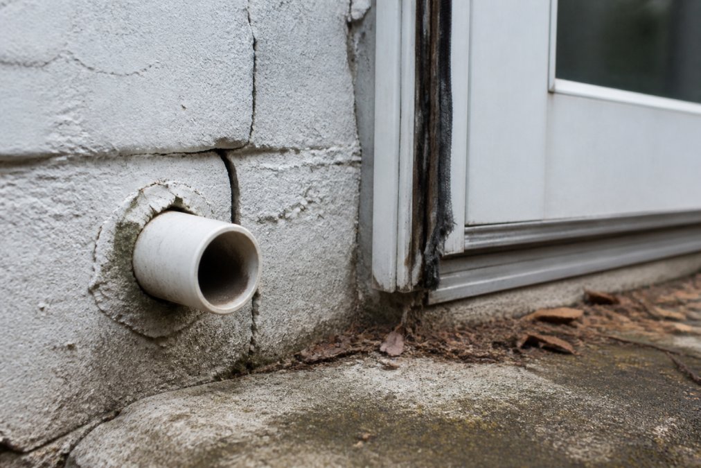 Close-up of a weathered home foundation showing a visible crack near a pipe entry point, with worn weatherstripping on a door frame in the background under overcast daylight