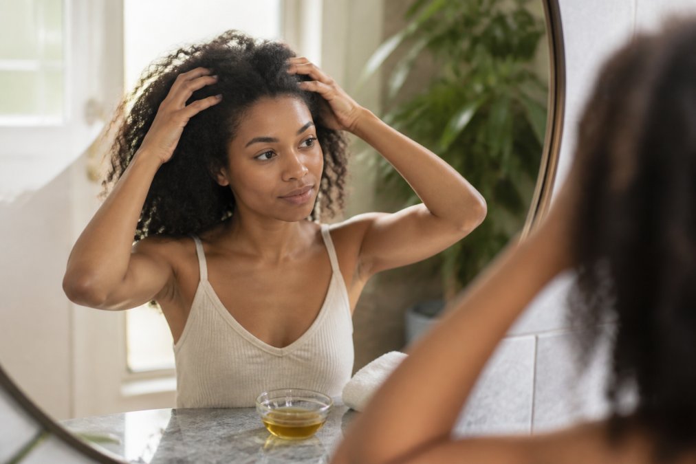 Woman with curly hair gently massaging her scalp with fingertips in a bright, sunlit bathroom.