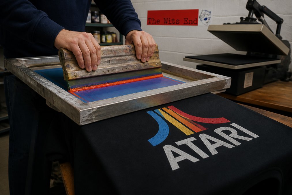 Close-up of a person's hands pulling a squeegee across a screen printing mesh onto a retro Atari graphic t-shirt in a small print shop, with colorful plastisol ink and a heat press visible in the background