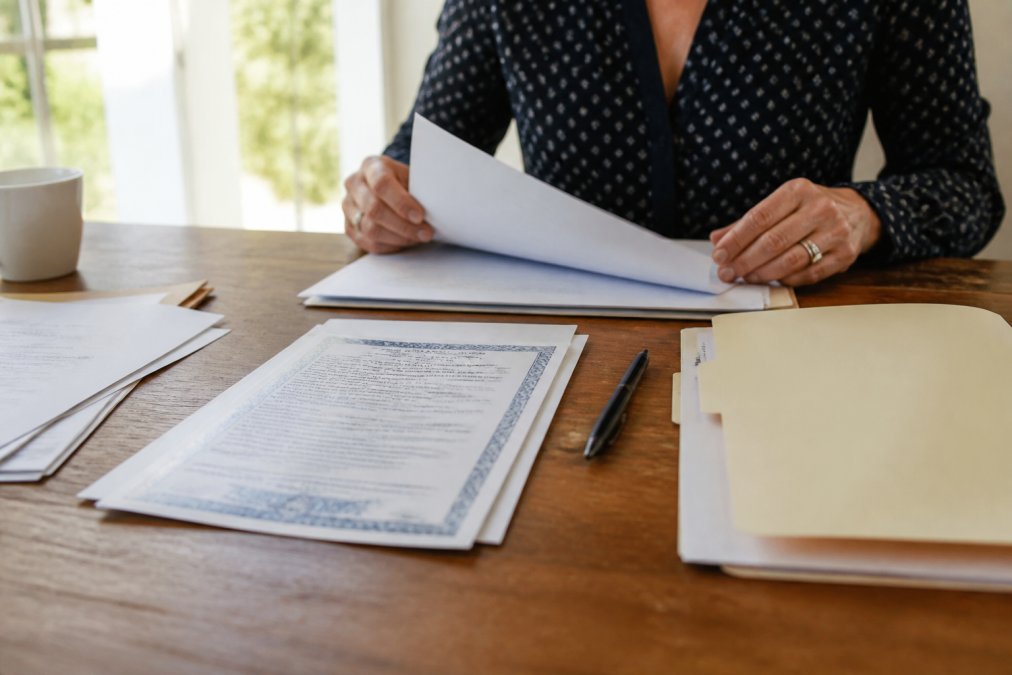 Person organizing documents and deed on desk with pen and folder in organized arrangement, preparing for real estate transaction with natural morning light