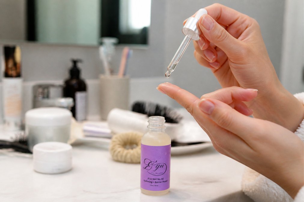 Close-up of hands applying a small drop of serum to fingertip before gently patting onto clean facial skin in natural bathroom lighting
