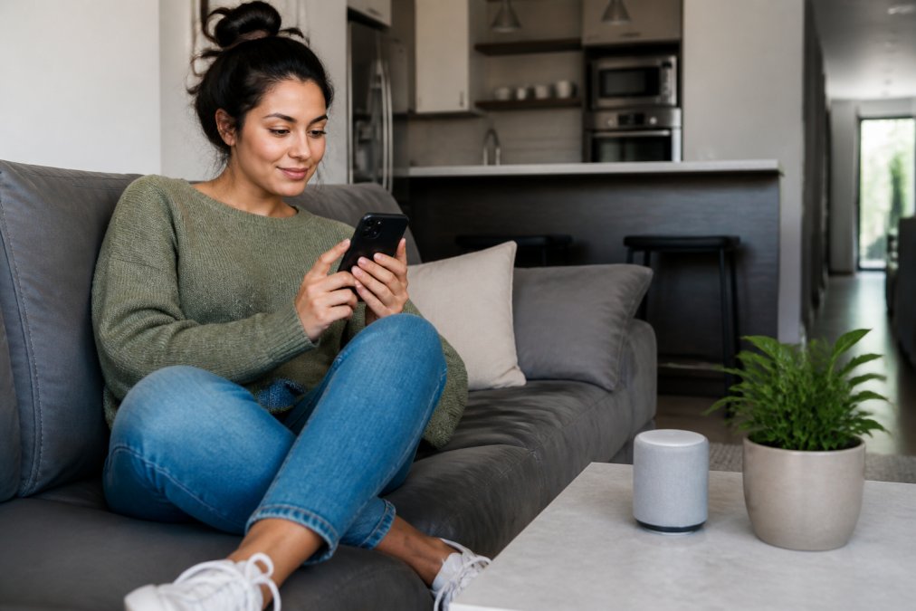 A young adult sitting on a couch scrolling a social feed on a smartphone while a small trending gadget sits on the coffee table beside them