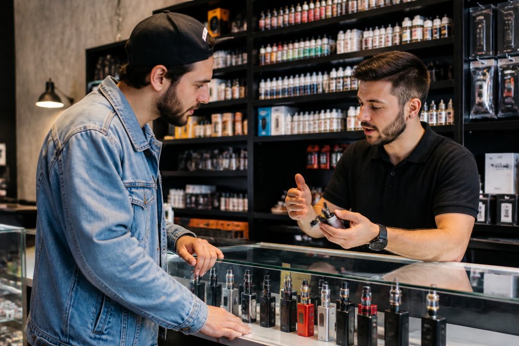 A knowledgeable vape shop staff member assisting a customer in a welcoming retail environment, discussing device options and answering questions face-to-face with premium brands visible on shelves