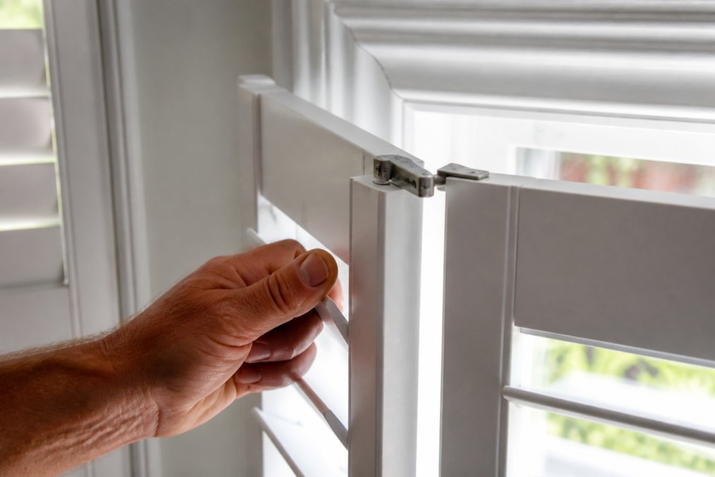 Close-up of a hand opening a white plantation shutter panel, revealing the top hinge and pivot points in bright natural light from a window