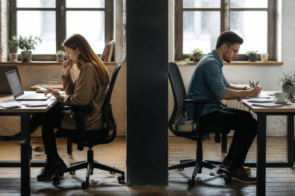 Two team members at separate desks facing away from each other, with laptops and documents scattered, symbolizing disconnected workflows and lack of collaboration