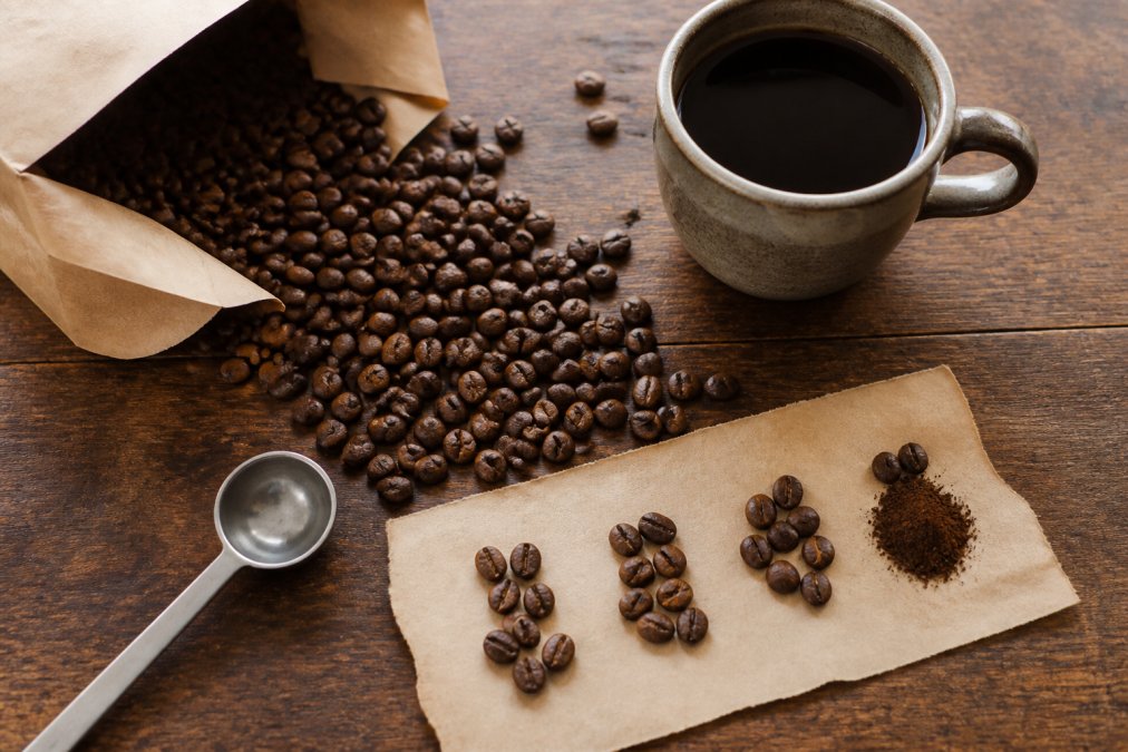 Overhead view of freshly roasted single origin coffee beans spilling from an open kraft paper bag with a ceramic cup of brewed coffee, tasting notes on paper, and a small metal cupping spoon on a wooden surface