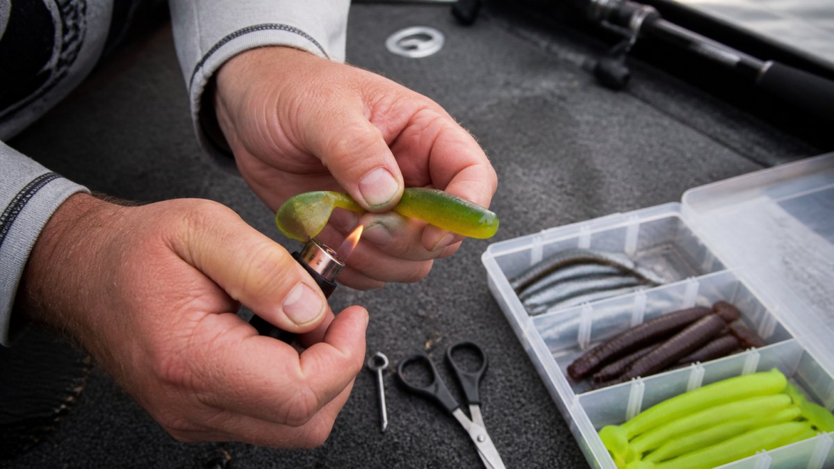 Close-up of an angler's hands on a boat deck holding a torn soft plastic swimbait and a lighter, about to heat-seal the split tail, tackle box open nearby