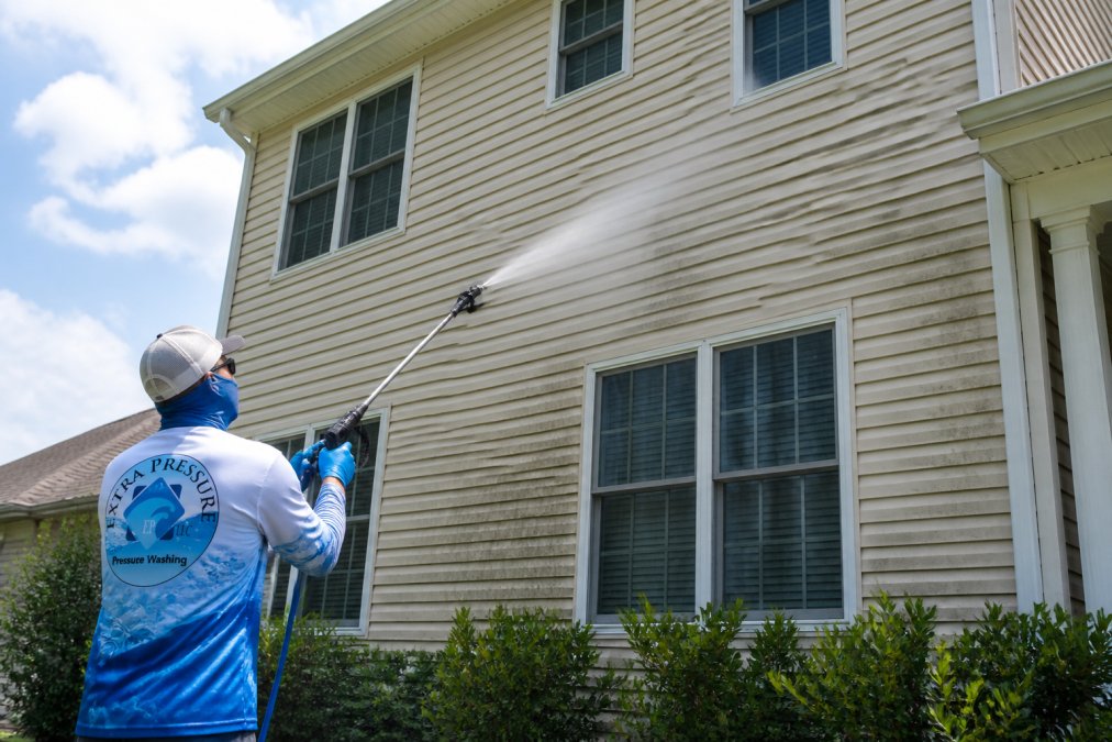 A professional technician in safety gear using a low-pressure sprayer to apply cleaning solution to a two-story home's vinyl siding, with visible soft mist and no harsh spray, on a sunny day