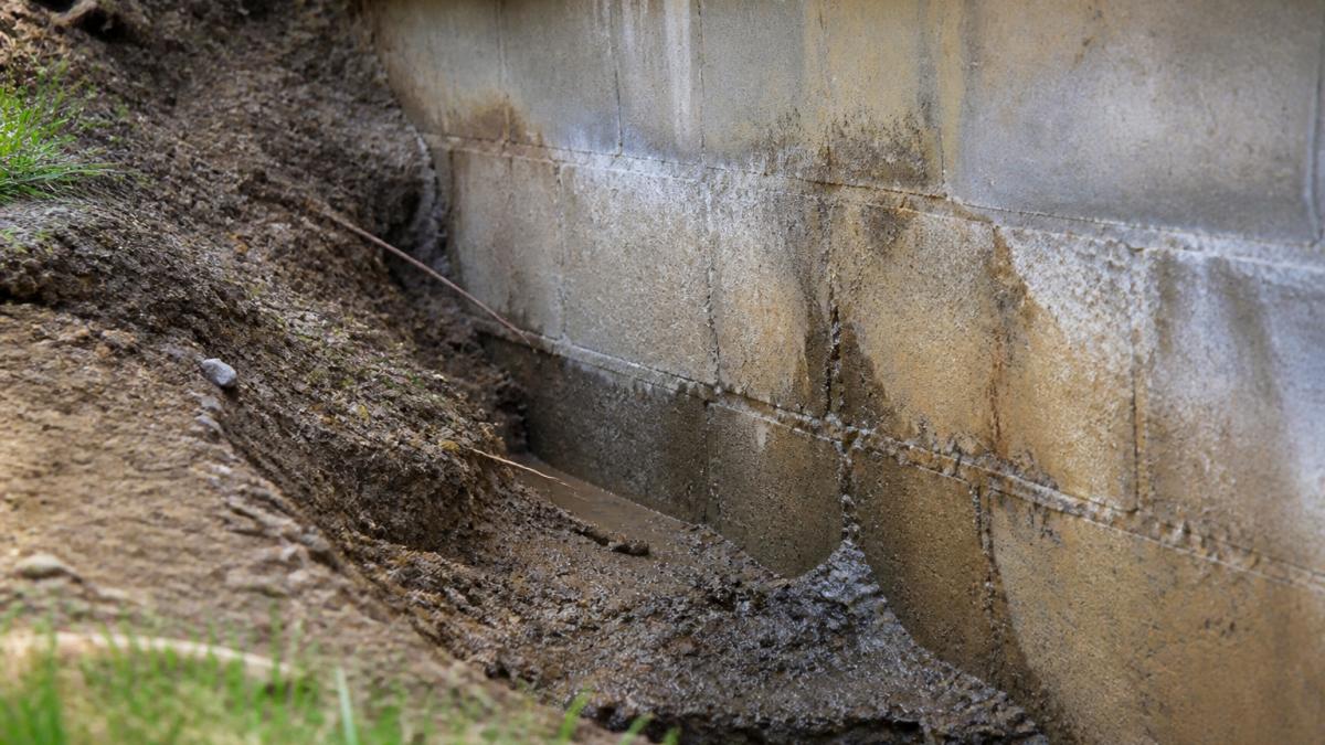 Close-up of exposed foundation wall with visible soil erosion, water staining, and areas where topsoil has washed away revealing bare earth