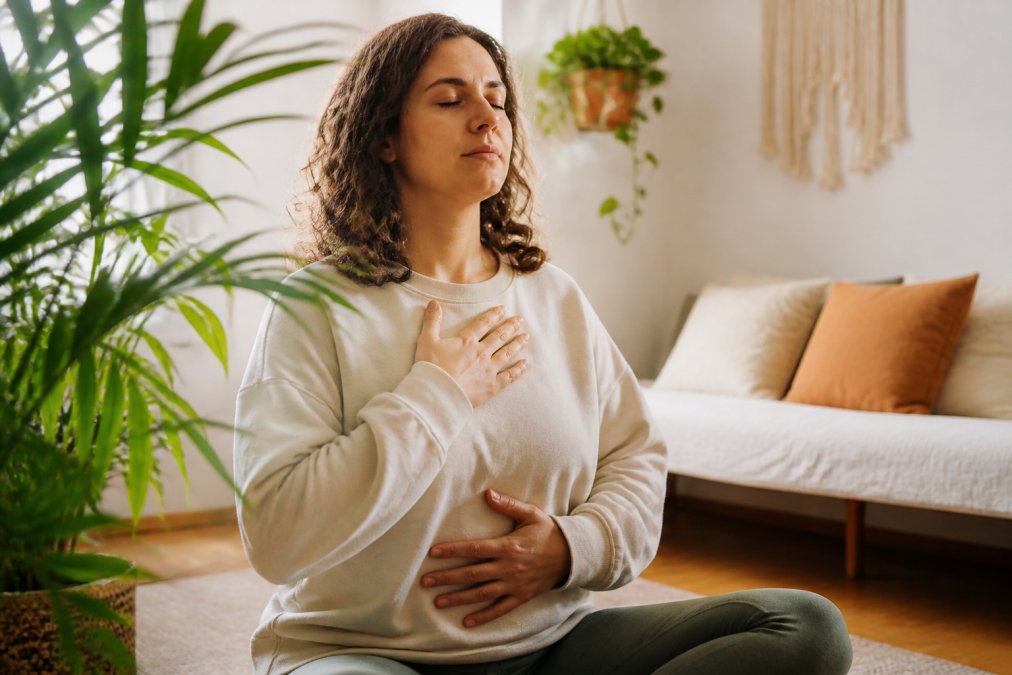A woman in comfortable clothing practicing deep breathing or meditation in a calm, sunlit room with plants, appearing relaxed and focused on her wellbeing