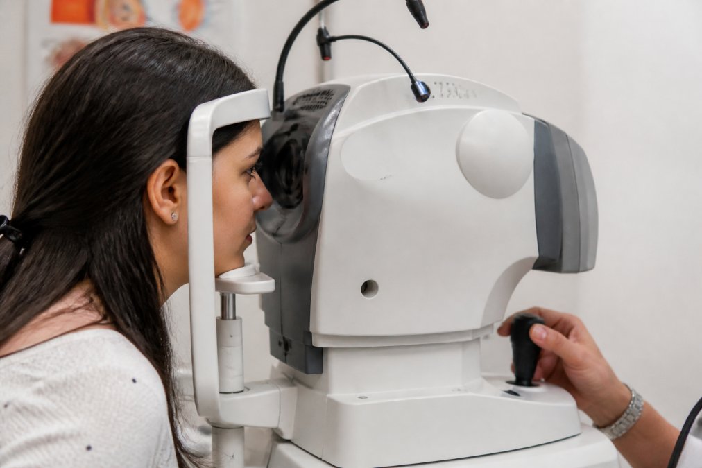 Close-up of advanced eye imaging equipment (OCT or retinal imaging device) with a patient positioned at the eyepiece during a diagnostic scan, showing the clinical setting with soft lighting and medical technology