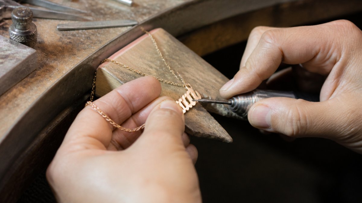 Close-up of artisan's hands carefully crafting a custom name necklace at a workbench, with polished metal, engraving tools, and materials visible under focused task lighting, showing precision and deliberate technique