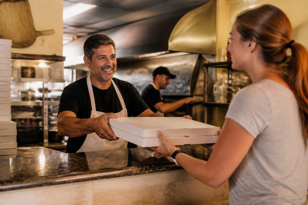 A pizzeria owner handing a pizza box across the counter to a smiling customer, with a busy open kitchen visible behind them