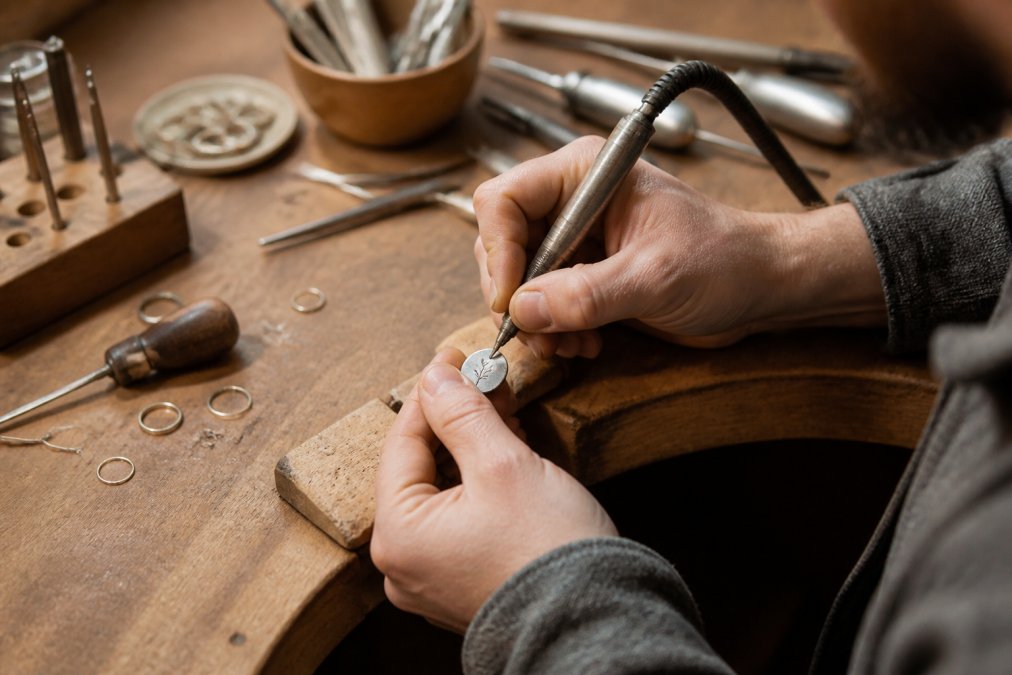 Close-up of artisan hands carefully crafting a personalized metal jewelry piece on a wooden workbench, with tools and materials visible in warm studio lighting