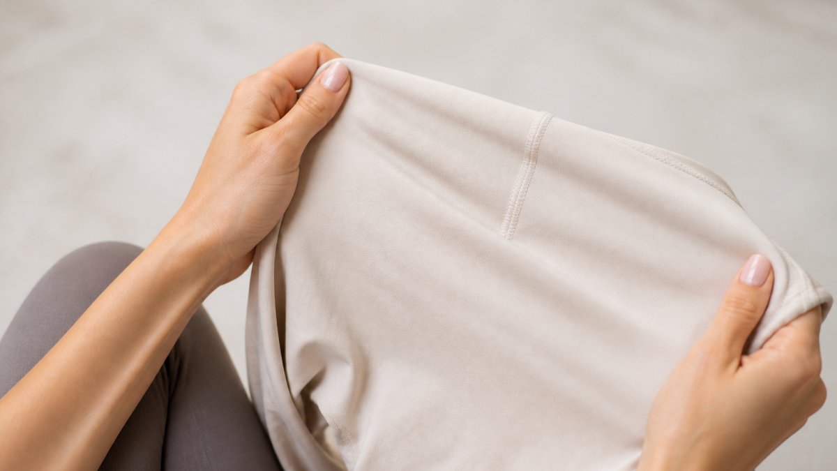 Close-up of a woman's hands holding soft, light-colored athletic leggings fabric against a bright neutral background, showing the texture and stretch of the material