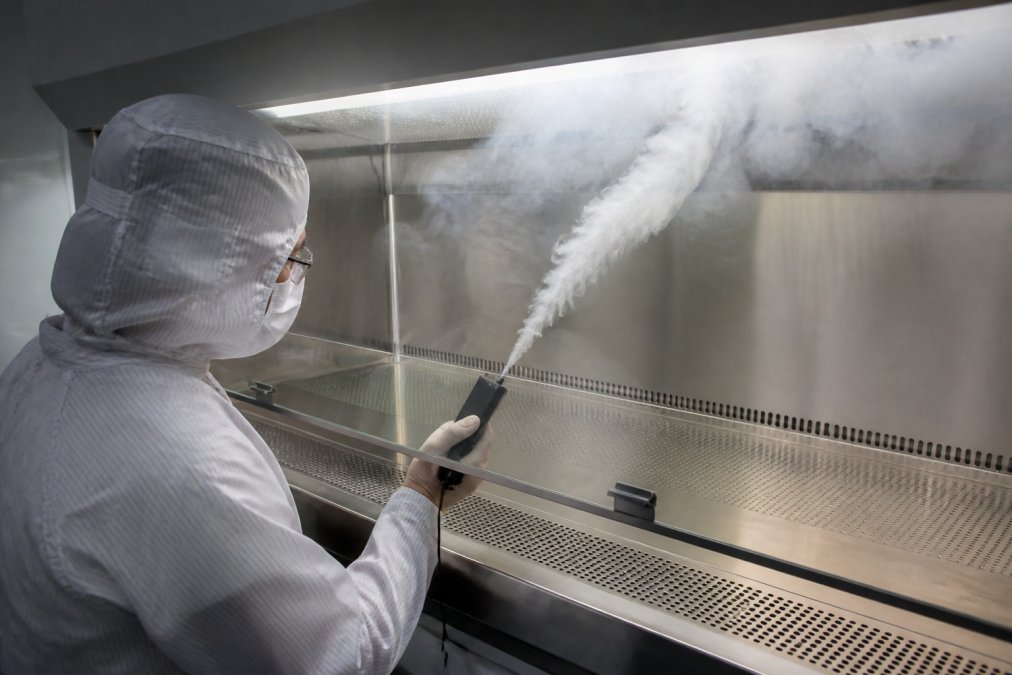 Cleanroom technician in protective equipment performing smoke testing inside a laminar flow hood, observing smoke particle behavior and airflow patterns in professional laboratory setting with controlled lighting