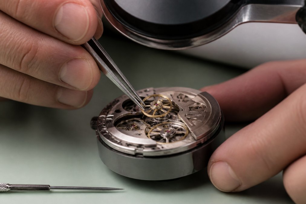 Close-up of a watchmaker's hands using precision tweezers to seat a balance wheel into an automatic movement under a magnifying lamp in a clean workshop