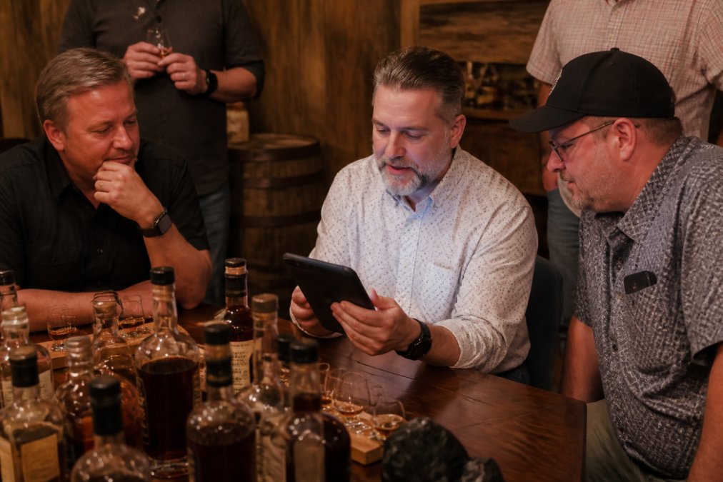 Group of bourbon collectors gathered at a tasting event, with one person showing their collection app on a tablet to others while bottles are arranged on a wooden table, warm lighting creating an intimate atmosphere