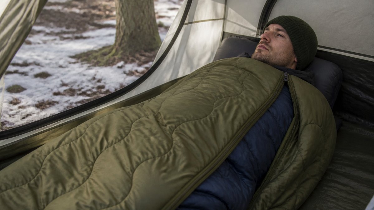 Camper in a tent demonstrating three-sided zipper configuration of a woobie draped over a sleeping bag during winter camping, showing proper headport positioning and layering technique with natural tent lighting