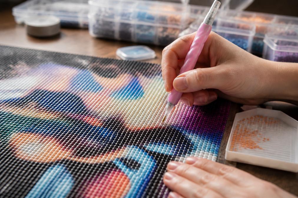 Close-up of a crafter's hands holding an ergonomic diamond painting pen with a cushioned grip, working on a vibrant full-drill canvas on a desk with organized trays nearby