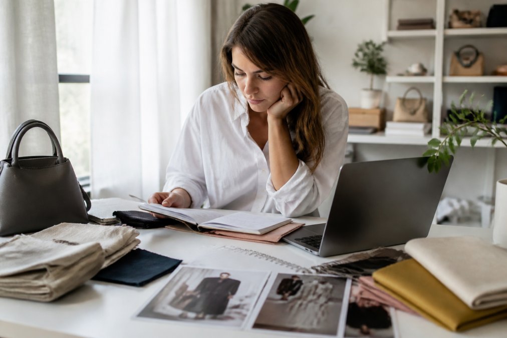Person sitting at a desk reviewing clothing items in a notebook with a laptop open, surrounded by fabric swatches and style inspiration images, natural afternoon lighting from a window