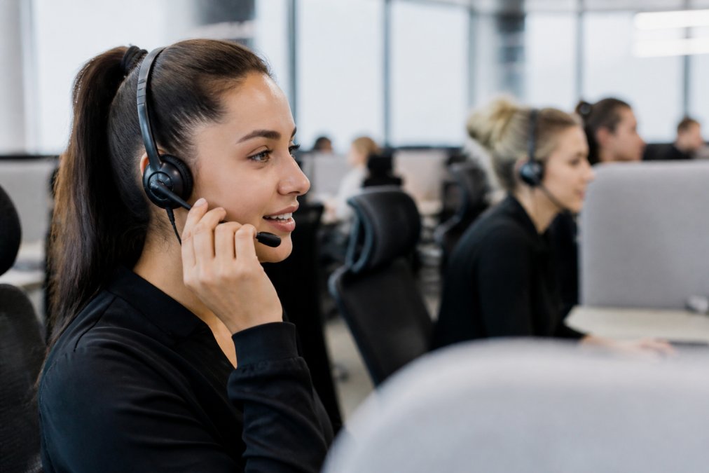 A calm support agent wearing a headset in a modern call centre, speaking reassuringly while a computer screen shows a vehicle dispatch map in the background