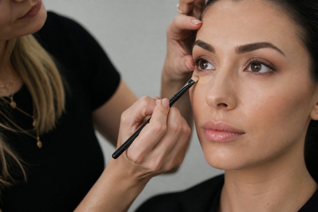 Professional makeup artist's hands using a precision angled brush to apply concealer with controlled, intentional strokes on a client's face, showing the bristle contact and blending technique